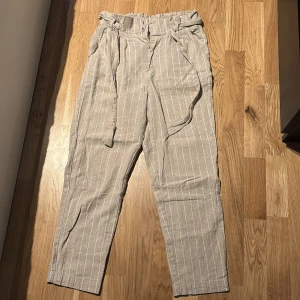 Beige randiga byxor  - Snygga beige tygbyxor från Gina Tricot med vita ränder, hög midja och knytskärp i midjan. Byxorna har avslappnad passform, fickor i sidorna och är perfekta för varmare dagar. Byxorna är i storlek 36. 
