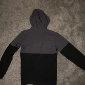 Under Armour svart/grå vindjacka - Snygg svart och mörkgrå vindjacka från Under Armour med huva och hel dragkedja. Jackan har lång ärm, bröstficka med dragkedja och logga på bröstet. Perfekt för dig som gillar sportig stil och vill ha något lätt och funktionellt.