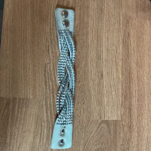 Flätat ljusgrönt mocka-armband med nitar - Snyggt armband i ljusgrön mocka med flätad design och glittriga silverfärgade nitar på ovansidan. Stängs med tryckknappar i silver. Perfekt accessoar för att lyfta din outfit med lite extra bling.