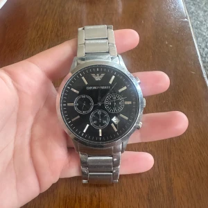Emporio Armani herrklocka i stål - Stilren klocka från Emporio Armani med svart urtavla, tre mindre visartavlor och datumvisning. Boett och länk i silverfärgat rostfritt stål. Klassisk rund form och tydliga indexmarkeringar. Perfekt accessoar för dig som gillar en clean och modern look.