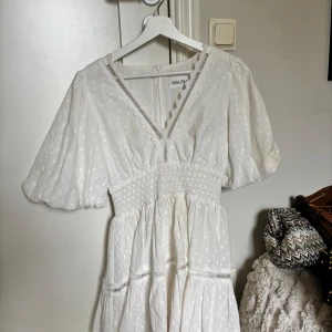 Vit kortklänning från Malina - Superfin vit kortklänning från Malina med puffärmar, v-ringning med spetsdetaljer och smockad midja. Klänningen har diskret prickigt mönster, volangdetaljer och dragkedja bak. Perfekt för sommarens alla tillfällen!