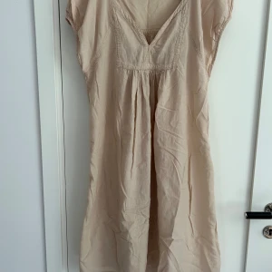 Beige kortklänning från Rabens Saloner - Supersöt beige kortklänning från Rabens Saloner med v-ringning och lätt puffade korta ärmar. Klänningen har en avslappnad passform och är gjord i mjuk bomull, perfekt för varma dagar. Fina sömdetaljer och rynk vid byst och rygg ger en bohemisk känsla.