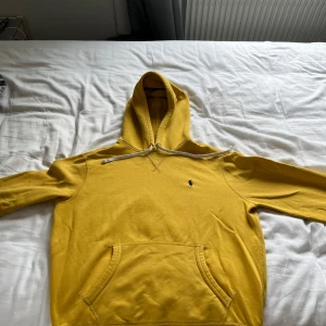 Gul hoodie från Polo Ralph Lauren - Gul hoodie från Polo Ralph Lauren med klassisk blå broderad logga på bröstet. Tröjan har huva med vit snörning och en stor magficka. Tillverkad i mjuk bomull, perfekt för chill dagar. Långärmad och normal passform.