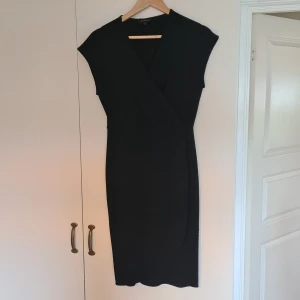 Svart omlottklänning Alice Bizous I storlek  S - Säljer en svart klänning från Alice Bizous i storlek S. Klänningen är knälång med v-ringning och ärmlös design. Tyget har en lätt glittrig finish som ger en snygg touch. Stretchigt och följsamt material.Perfekt för dig som gillar stilrena och eleganta plagg.
