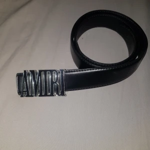 Svart bälte med AMIRI-spänne - Snyggt svart bälte från AMIRI med ett stort silverspänne där märkesnamnet står ut. Bältet är gjort i skinn och har en klassisk, stilren design som passar till jeans eller kostymbyxor. Perfekt accessoar för att lyfta din outfit.