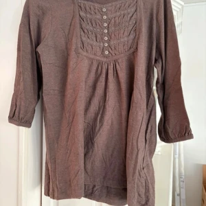 Brun långärmad blus från Isolde - Säljer en brun långärmad blus från Isolde med dekorativa knappar och veckad detalj framtill. Blusen har trekvartsärm och är tillverkad i mjukt bomullsmaterial. Perfekt för en avslappnad och stilren look.