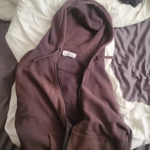 Brun hoodie från Stone Island - Säljer en brun hoodie från Stone Island med klassisk logga på ärmen. Tröjan har dragkedja och huva, perfekt för en avslappnad stil. Mjuk insida och två fickor framtill.