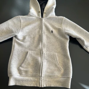 Ljusgrå hoodie från Polo Ralph Lauren - Säljer en ljusgrå hoodie från Polo Ralph Lauren med dragkedja och huva. Tröjan har ribbad struktur, två fickor framtill och den klassiska broderade loggan på bröstet. Perfekt för en avslappnad stil. Tar även imot byten!