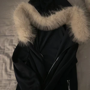 Svart jacka med fuskpäls och dödskalle - Svart jacka med stor huva kantad av ljus fuskpäls. Jackan har dragkedja framtill och ett broderat dödskallemotiv i rosa på bröstet. Snygg och edgy stil, perfekt för dig som vill sticka ut.