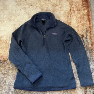 Mörkblå fleece-tröja från Patagonia - Säljer en mörkblå fleece-tröja från Patagonia med halv dragkedja och hög krage. Tröjan har lång ärm och en praktisk ficka med dragkedja på ena ärmen. Perfekt för kyliga dagar och outdoor-aktiviteter. Lite nopprig men det går att fixa, sen ett litet hål som man knappt märker av eller ser. 