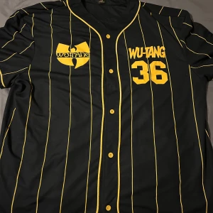 Wu-Tang Clan svart baseballskjorta med gula ränder - Svart baseballinspirerad skjorta från Wu-Tang Clan med gula vertikala ränder, tryckt logga och texten 'WU-TANG 36' fram samt stor logga och 'Enter The 36 Chambers' bak. Knappar framtill och klassisk basebollkrage.