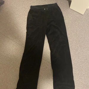 Svarta mjukisbyxor från Juicy Couture - Svarta mjukisbyxor från Juicy Couture med resår och öljetter i midjan. Byxorna har raka ben och bakfickor med knappar. Perfekta för en avslappnad stil. Saknas dock snören.