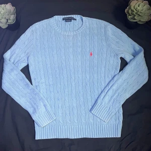 Ljusblå stickad tröja från Ralph Lauren - Säljer en ljusblå stickad tröja från Ralph Lauren med klassiskt kabelstickat mönster och rund halsringning. Tröjan har lång ärm och en liten rosa broderad logga på bröstet. Perfekt för lager-på-lager och stilrena outfits.