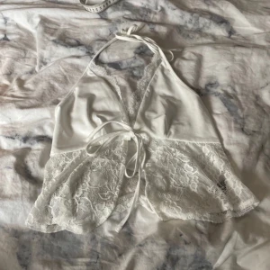 Vit spetsig halterneck topp med knytband - Säljer en vit halterneck topp med v-ringning och knytband i midjan. Toppen har spetsdetaljer längs urringningen och nertill, vilket ger en romantisk känsla. Perfekt för dig som gillar feminina detaljer och lätt material.