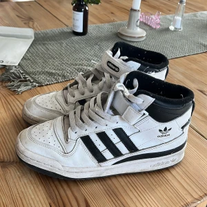 Vita och svarta Adidas sneakers - Säljer ett par klassiska Adidas sneakers i vitt med svarta detaljer och högt skaft. Jag säljer då de inte kommer till användning längre men de är använda. De är smutsiga men det är nog inget som inte går bort om man förökar tvätta de:)