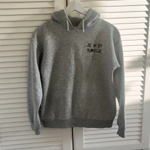 Grå hoodie med tryck 'JE M'EN AMOUR' - Säljer en grå hoodie med huva och tryck både fram och bak där det står 'JE M'EN AMOUR' samt en stjärna. Tröjan har ribbade muddar och en klassisk passform. Perfekt för en avslappnad stil.