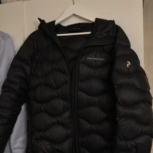 Svart/Mörkblå Peak Performance Helium jacka - Säljer en svart/mörkblå dunjacka från Peak Performance med huva och dragkedja. Jackan har en puffig design och logga på bröstet. Perfekt för kyliga dagar och riktigt skön att ha på sig.