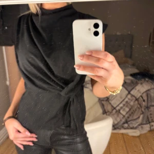 Svart knytblus - Snygg svart blus med hög hals och knyt i midjan. Blusen har en elegant drapering framtill och är ärmlös. Perfekt för att lyfta vilken outfit som helst.