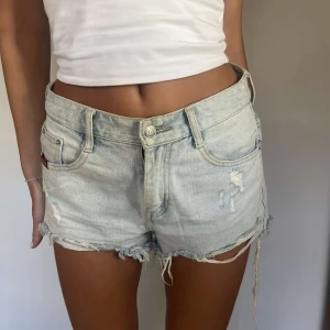 Ljusblå jeansshorts med fransar - Säljer ett par ljusblå jeansshorts med fransiga benslut och slitna detaljer både fram och bak. Klassisk femficksmodell med knapp och dragkedja. Perfekta för varma dagar och en avslappnad stil.