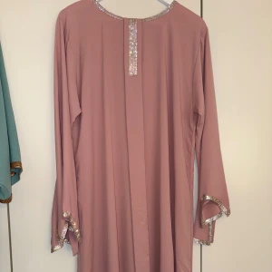 Lång rosa klänning med paljettkant - Säljer en lång, ljusrosa klänning med raka långa ärmar. Klänningen har dekorativa paljettdetaljer längs halsringningen, ärmsluten och framtill. Perfekt för dig som gillar enkel men glittrig stil. (Marockans Klänning) Köpt i Dubai. 