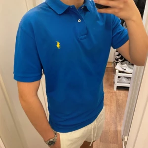 Polo Ralph Lauren (Blå piké) - Säljer en klassisk blå piké t-shirt från Polo Ralph Lauren med gul broderad logga på bröstet. Den är oanvänd och redo för en ny ägare. Perfekt för en stilren och sportig look. Pikén är size M!