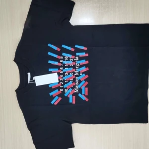 Svart t-shirt från Maison Margiela med grafiskt tryck - Svart t-shirt från Maison Margiela med ett färgstarkt grafiskt tryck i blått och rött samt vit text och siffror på bröstet. Klassisk passform och rund halsringning. Perfekt för dig som gillar statement-plagg.