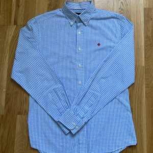 Blårutig skjorta från Morris - Snygg blårutig skjorta från Morris med klassisk krage och knappar framtill. Skjortan har ett broderat rött emblem på bröstet och är tillverkad i bomull. Perfekt för dig som gillar stilrena och tidlösa plagg. Sjortan är i väldigt bra skick. 