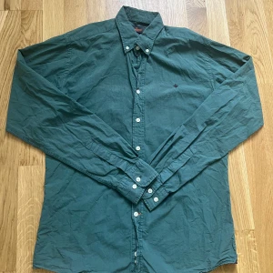 Grön skjorta från Morris - Snygg grön skjorta från Morris med klassisk krage och vita knappar. Skjortan har lång ärm och en liten broderad detalj på bröstet. Perfekt för dig som gillar stilrena plagg med diskreta detaljer. Skjortan är i väldigt bra skick men kan behöva strykas 