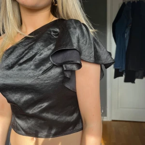 Svart oneshoulder topp med volang - Säljer en svart oneshoulder topp med snygg volangdetalj på axeln. Toppen har en glansig finish skriv för fler bilder eller frågor💞💞