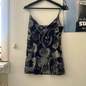 Svart och vit blommig topp från Lindex - Säljer en svart och vit topp från Lindex med blommigt mönster och tunna axelband. Toppen har en lätt utsvängd passform och v-ringning framtill. Perfekt för varma dagar! M-L