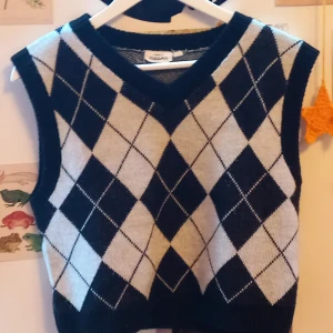 Svart och vit rutig stickad väst - Snygg stickad väst med svart och vit argylemönster och v-ringad hals. Perfekt att bära över en skjorta eller t-shirt för en trendig lager-på-lager-look.