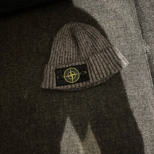 Grå ribbad mössa från Stone Island - Säljer en grå ribbstickad mössa från Stone Island med den klassiska logotypen framtill. Perfekt för kyliga dagar och enkel att matcha med olika outfits.