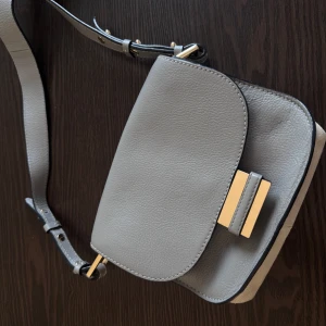 HM Ljusgrå axelväska med gulddetaljer - Säljer en stilren ljusgrå axelväska i skinn med justerbart axelband och snygga guldfärgade detaljer. Väskan har ett huvudfack och ett mindre innerfack, perfekt för det viktigaste. Enkel och modern design som passar till många outfits.