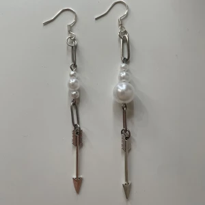 Silvriga örhängen med pärlor och pilhänge - Unika örhängen i silverfärg med vita pärlor och hängande pil-detalj. Snygg kombination av metall och pärlor som ger ett coolt och trendigt uttryck. Perfekta för att lyfta din stil med en personlig touch.