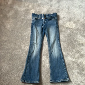 Blå bootcut jeans med bälte från H&M - Säljer ett par blå bootcut jeans från H&M med låg midja och justerbar midja. Jeansen har ett matchande jeansbälte som sitter fast på byxorna med silverfärgat spänne och klassiska fickor både fram och bak. Perfekta för en avslappnad och trendig look.