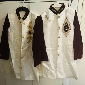 Vit och vinröd broderad lång kavaj med pärldetaljer som passar barn i ålder 8-10 år. 100 kr per st - Säljer en unik lång kavaj som passar barn i ålder 8-10 år med vit brokad och vinröda ärmar och krage. Utsmyckad med guldiga och pärlbroderade detaljer samt dekorativa knappar. Perfekt för festliga tillfällen där du vill sticka ut.