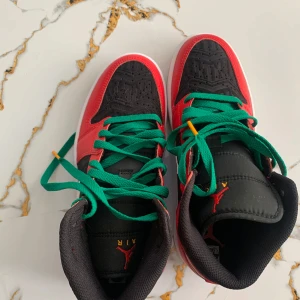 Nike Air Jordans med röd och svart färg - Säljer ett par Nike Air Jordans med svart tå och ovandel, röda paneler och vit swoosh. Skorna har gröna skosnören och klassisk Air Jordan-logga på plösen. Perfekta för dig som vill sticka ut med färg på fötterna.