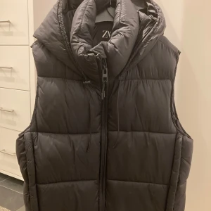 Svart dunväst med huva från Zara - Svart dunväst från Zara med vadderad huva och dragkedja framtill. Enkel att ha med olika outfits, både sommar och vinter. Västen är vattenavvisande, vindtät, lättviktig och andas.
