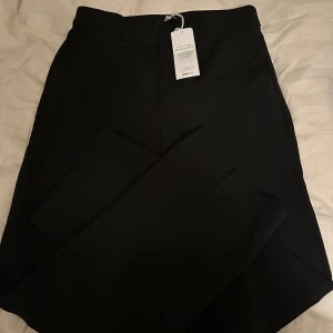 Svarta kostymbyxor i rak modell - Säljer ett par klassiska svarta kostymbyxor med normal passform och raka ben. Byxorna har hällor för bälte och stängs med dragkedja och knapp och har även en hög midja . Perfekta till en stilren look.