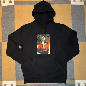 Svart hoodie med tryck - Säljer en svart hoodie med en färgglad tryckt bild framtill som föreställer en person vid en röd bil. Klassisk huva och känguruficka. Perfekt för en avslappnad stil.                                                                                     Helt ny✅.                                                                                          Storlek: L finns olika storlekar.                                                      Rabatt om man följer 
