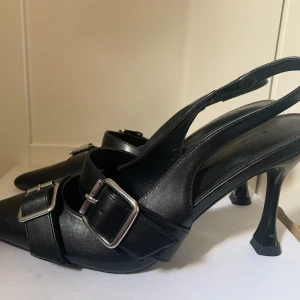 Svarta pumps med spänne och klack - Säljer ett par svarta pumps med spetsig tå och öppen häl. Skorna har två breda remmar med stora silverspännen och en hög, smal klack. Tillverkade i skinnliknande material och passar perfekt för dig som gillar en edgy och stilren look. Har aldrig används.