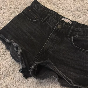 Svarta jeansshorts från Gina Tricot - Snygga svarta jeansshorts från Gina Tricot med fransiga benslut och klassisk femficksdesign. Shortsen har knapp och dragkedja framtill samt råa kanter för en cool look.