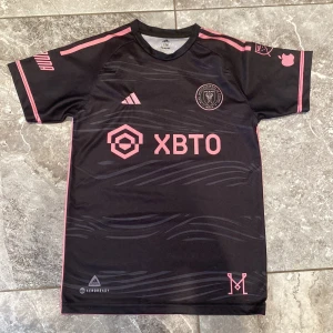 Inter Miami CF Messi 10 fotbollströja - Snygg svart Inter Miami CF fotbollströja med rosa detaljer och Messi 10 på ryggen. Kortärmad modell med klubbmärke och sponsorlogga på bröstet. Tillverkad i lätt och ventilerande material, perfekt för match eller träning.