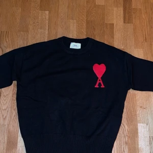 Svart stickad tröja med rött hjärta från Ami Paris - Svart stickad tröja från Ami Paris med ett stort rött hjärta och bokstaven A broderat på bröstet. Tröjan har rund halsringning och långa ärmar. Perfekt för dig som gillar stilrena plagg med en twist.