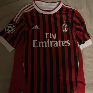 AC Milan Zlatan Ibrahimović 11 matchtröja - Säljer en klassisk AC Milan fotbollströja från Adidas med Zlatan Ibrahimović och nummer 11 på ryggen. Tröjan är röd och svart med vita detaljer, randigt mönster och Champions League-märke på ärmen. Perfekt för fans av Milan och Zlatan!