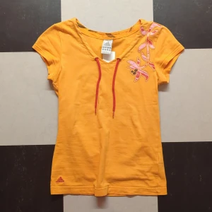 Gul t-shirt från Adidas Y2K  - Säljer en gul t-shirt från Adidas med röd broderad logga nertill och dekorativt med blommor och snörning vid halsen. T-shirten har korta ärmar och är tillverkad i mjuk bomull. Perfekt för en färgglad och sportig look!