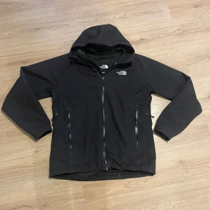 Svart vindjacka från The North Face - Säljer en svart vindjacka från The North Face med huva och dragkedja framtill. Jackan har två sidofickor med dragkedjor och är perfekt för blåsiga dagar. Den är stilren och funktionell, idealisk för utomhusaktiviteter. Bara att skriva om det är något man funderar på. 