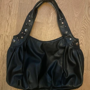Svart axelväska med nitar - Säljer en svart axelväska med coola nitar på handtagen. Väskan har en rymlig insida och en dragkedjeficka på baksidan. A4 får plats i väskan. Lägg märke till att den är lite sliten på banden och insidan, men det går att fixa. 