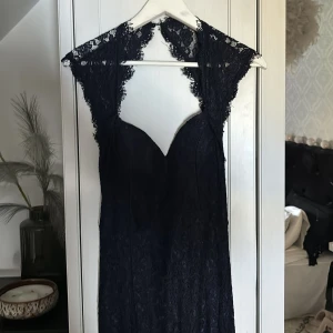 Mörkblå långklänning med spets och öppen rygg - Elegant svart långklänning med spetsdetaljer och djup v-ringning. Klänningen har öppen rygg och fina spetskanter vid axlarna. Perfekt för festliga tillfällen när du vill sticka ut med en sofistikerad look.