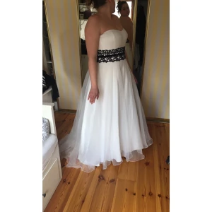 NY! Vit brudklänning med svart spetsdetalj - NY!                                                       Klänningen har en vacker, utsvängd kjol i flera lager och markerad midja med dekorativ spetsdetalj i svart.  Perfekt för bröllop, bal eller fest. Klänningen är oanvänd med lapparna kvar, förvarats väl i den tillhörande klädpåsen. Finns tunna band till, i klänningen. Jag hittar ingen stl lapp i klänningen eller på påsen men skulle säga att den är stl42 ungefär. Jag hade ungefär den stl när det var tänkt att klänningen skulle ha används. 